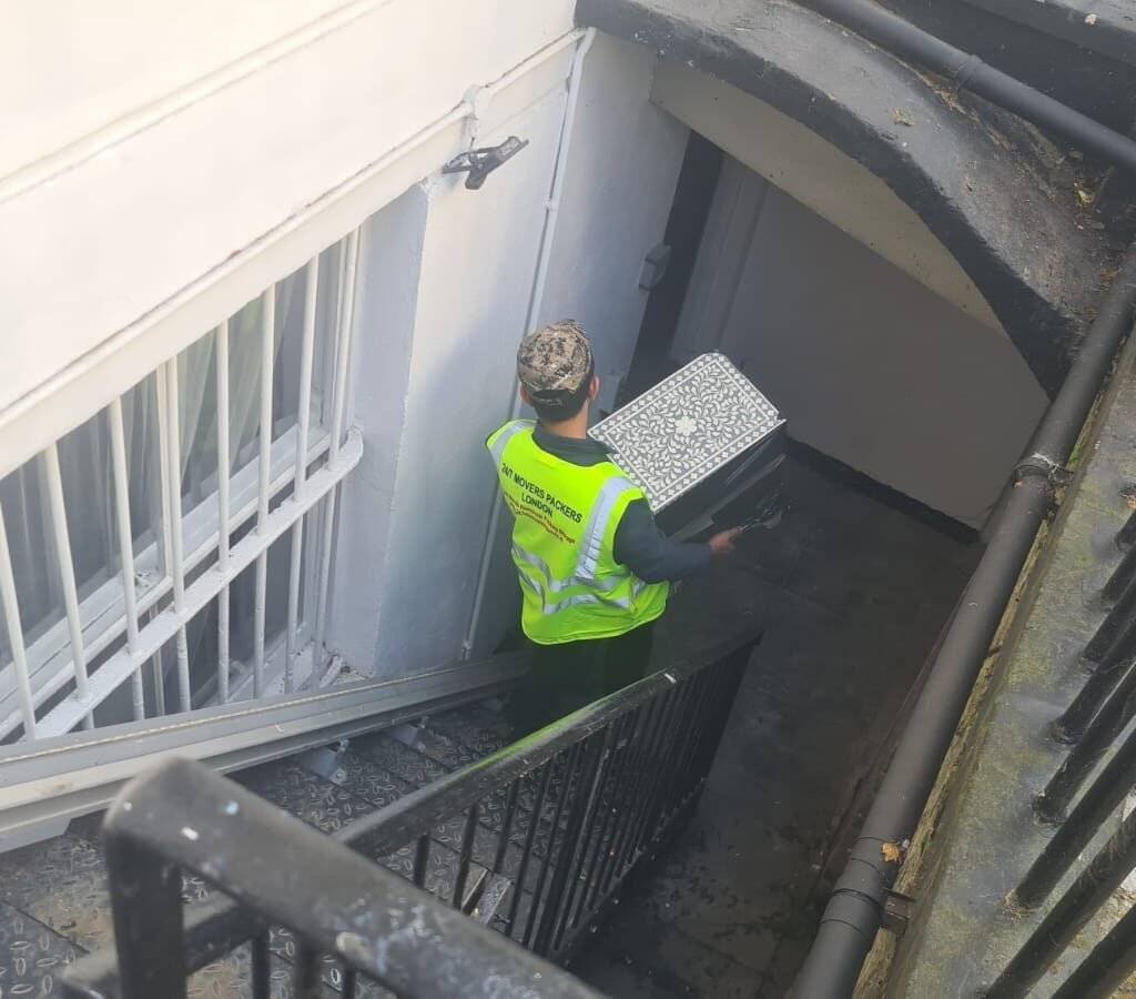 A mover wearing a high-visibility vest carries a patterned box down a staircase into a basement entrance.