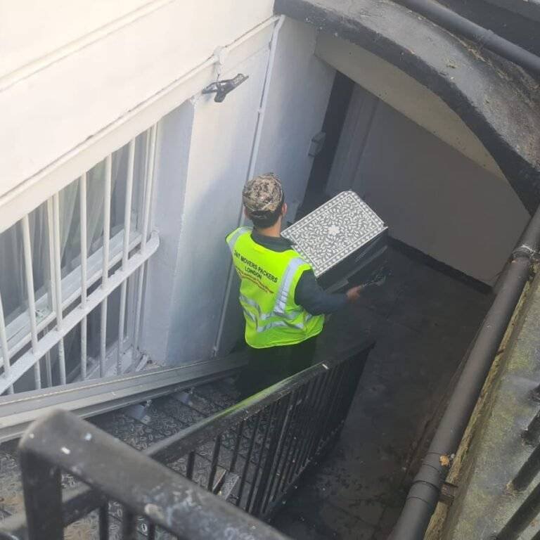 A mover wearing a high-visibility vest carries a patterned box down a staircase into a basement entrance.