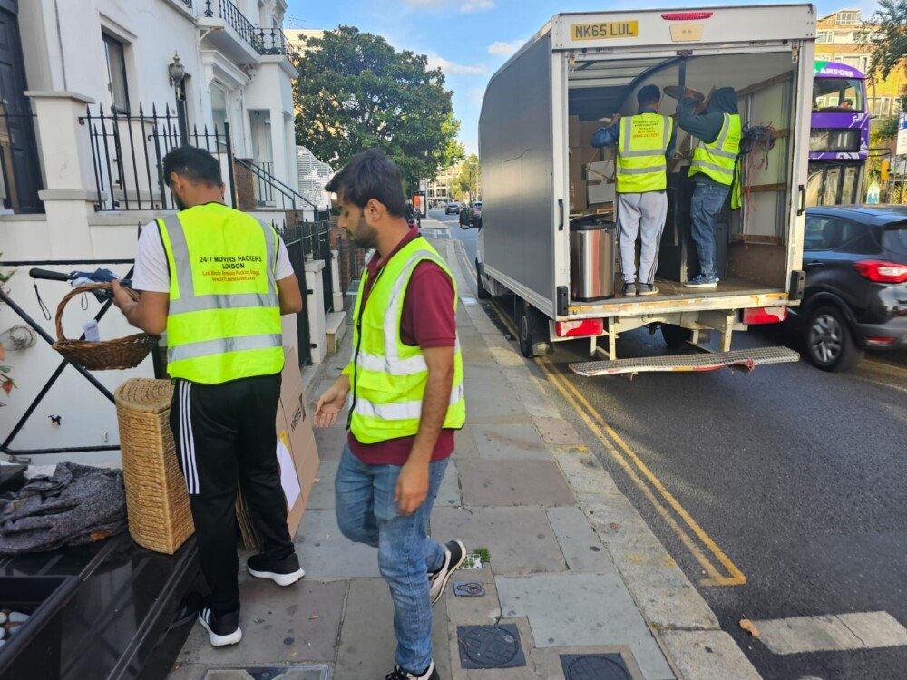 A mover wearing a high-visibility vest loads cardboard boxes into the back of a moving truck parked on a street with "NO PARKING" painted on the ground.