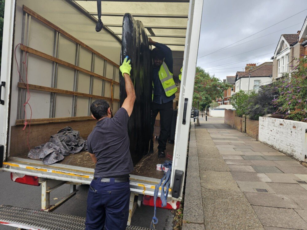 Two movers, one inside and one outside a moving truck, load a large black item while parked on a residential street.