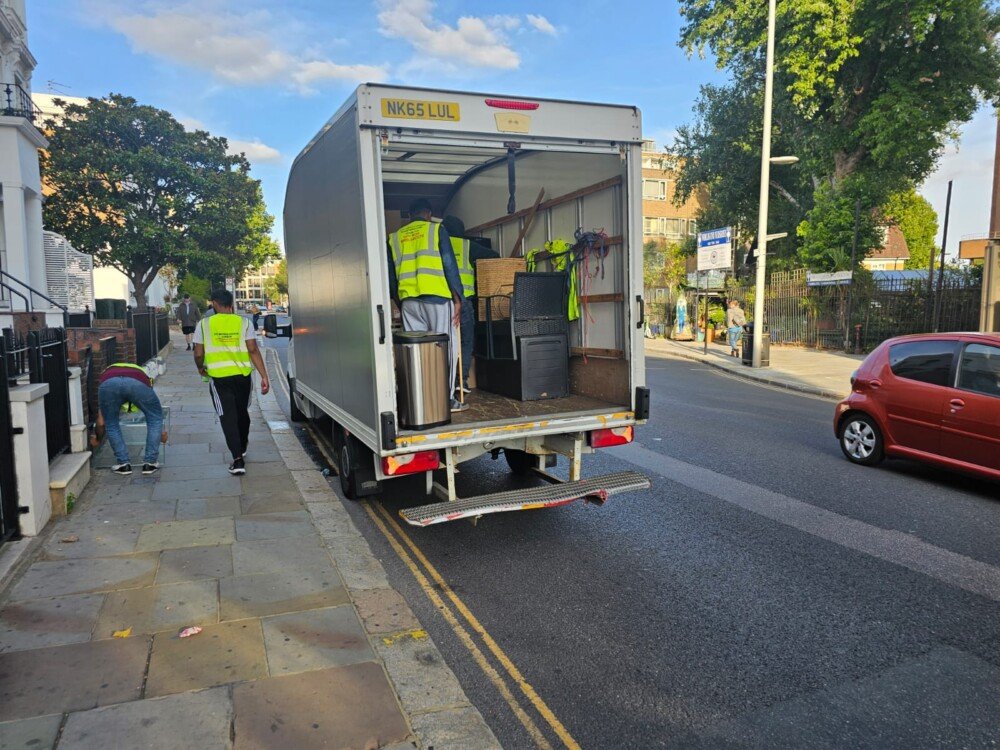 Movers in high-visibility vests unload items, including furniture, from the back of a moving truck parked on a residential street.
