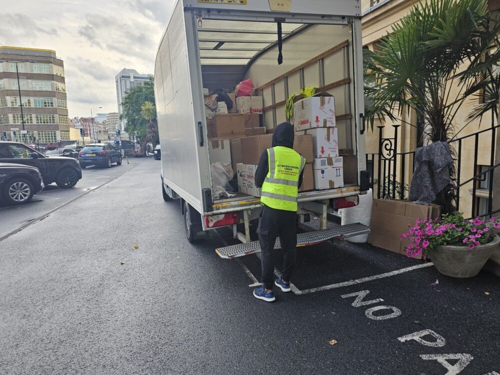 A mover wearing a high-visibility vest loads cardboard boxes into the back of a moving truck parked on a street with "NO PARKING" painted on the ground.