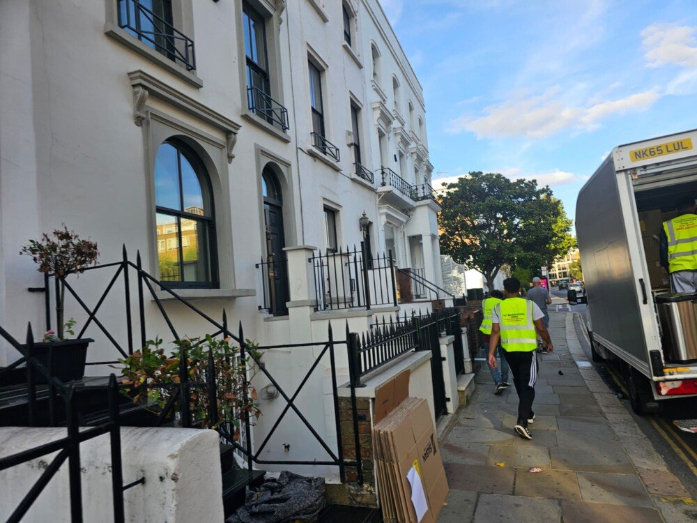 Several movers in high-visibility vests load household items into a moving truck outside a row of white townhouses.